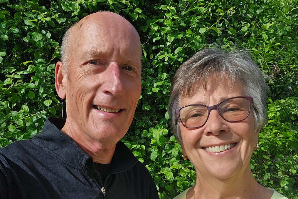 A smiling older couple stands outdoors in bright sunlight with lush green trees and shrubs behind them. The man, wearing a black quarter-zip pullover, and the woman, wearing purple-framed glasses and a light green top, are standing close together and looking at the camera. The photo is a close-up selfie with a vibrant, leafy background.