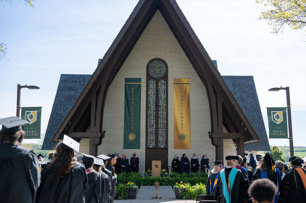 Keuka College graduates and faculty wearing regalia from behind standing in front of Norton Chapel