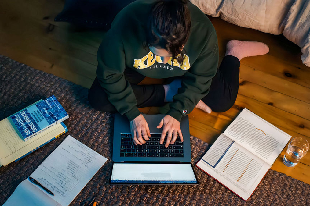 A student wearing a green Keuka College sweatshirt sits cross-legged on a wooden floor, typing on a laptop while surrounded by open textbooks, handwritten notes, a notebook, and a glass of water. The overhead view highlights a focused late-night study session in a cozy room.