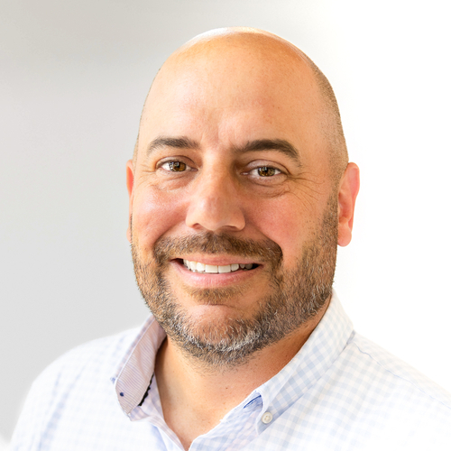 A smiling man with a shaved head and neatly trimmed beard wears a light blue button-down shirt in a professional headshot. He faces the camera against a softly blurred light-colored background.
