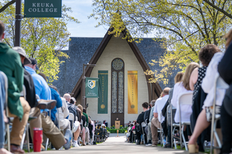 Wide view of Keuka College Commencement ceremony with graduates, families, and faculty seated outdoors facing the campus chapel stage on a sunny spring day.