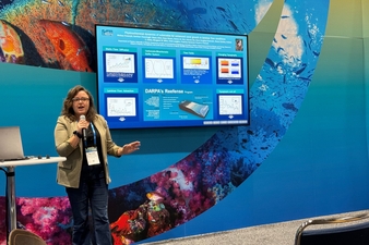 A person stands at the front of a conference presentation area, speaking into a microphone and gesturing toward a large digital poster displayed on a screen behind them. The presentation appears to focus on coral reef research, with charts, graphs, and diagrams visible on the screen. The backdrop features colorful underwater imagery, including coral reefs and fish, creating an ocean-themed setting. A laptop sits on a nearby table, and audience seating is visible in the foreground.