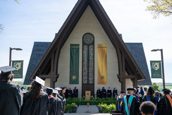 Keuka College graduates and faculty wearing regalia from behind standing in front of Norton Chapel