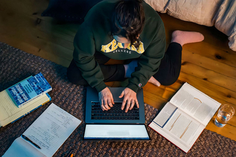 A student wearing a green Keuka College sweatshirt sits cross-legged on a wooden floor, typing on a laptop while surrounded by open textbooks, handwritten notes, a notebook, and a glass of water. The overhead view highlights a focused late-night study session in a cozy room.
